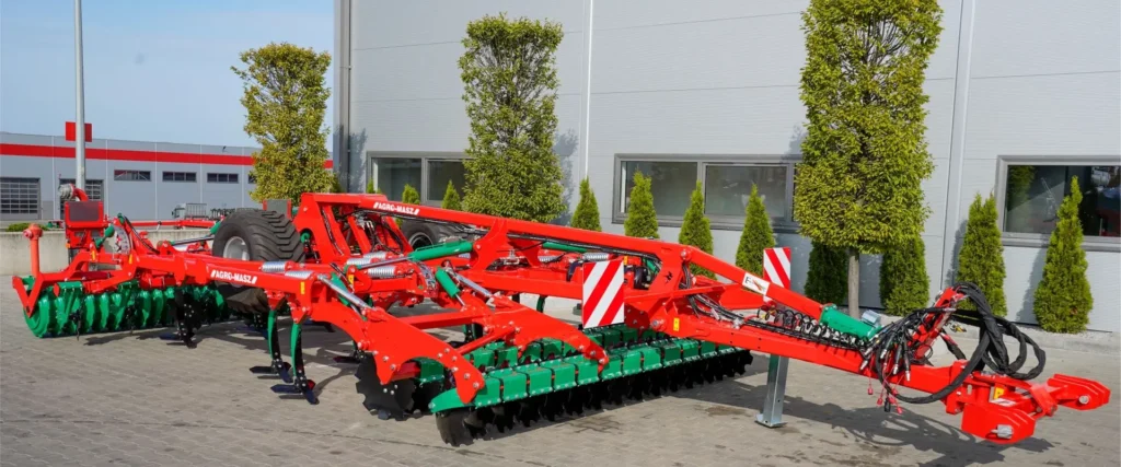 Zbliżenie na dużą, nowoczesną maszynę rolniczą marki Agro-Masz w kolorach czerwonym i zielonym, zaparkowaną na placu z kostki brukowej przed budynkiem firmy. Jest to zaawansowany agregat uprawowy lub siewnik z widocznymi rzędami talerzy tnących, hydraulicznymi siłownikami oraz dużymi kołami transportowymi. W tle widać nowoczesną halę przemysłową oraz ozdobne krzewy.