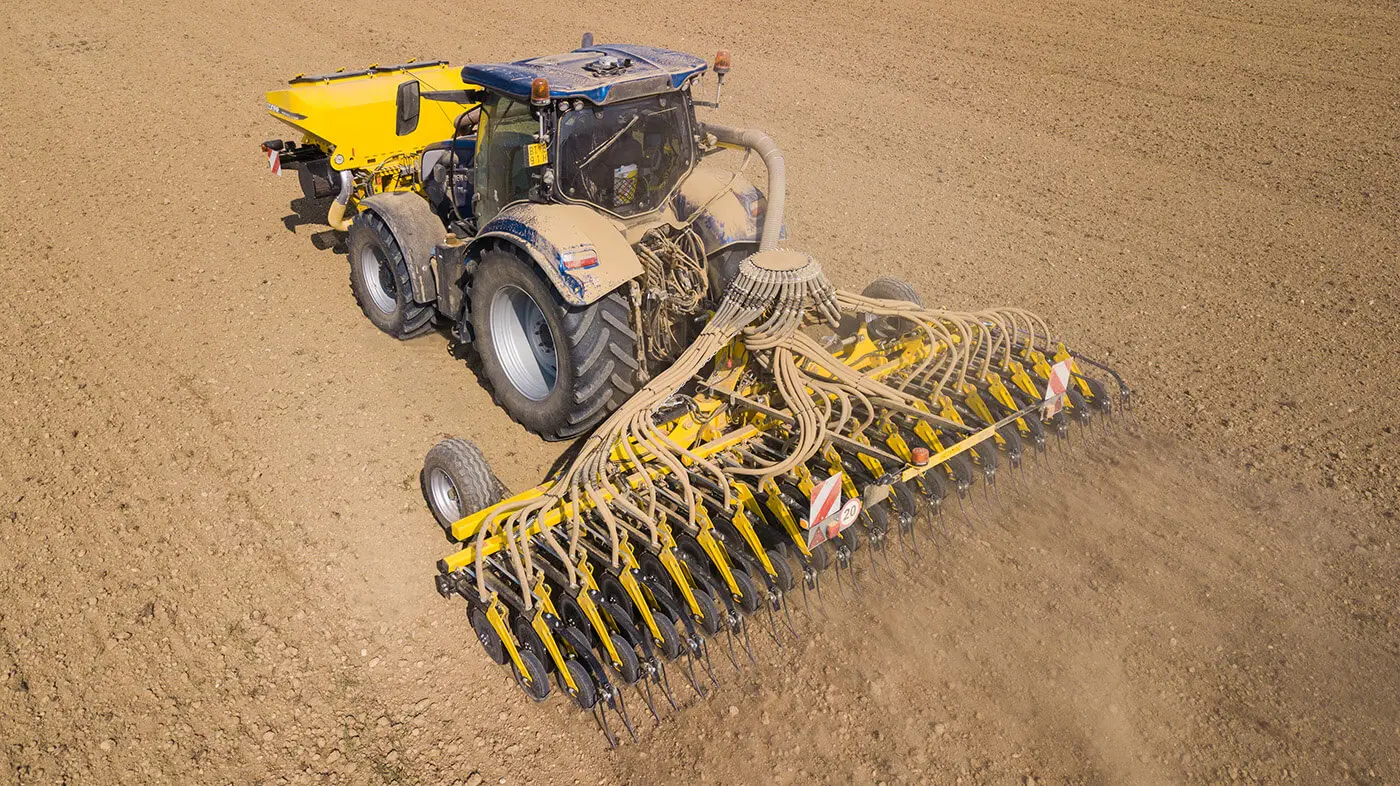 Widok z góry na niebieski ciągnik rolniczy pracujący w polu z żółtym siewnikiem zbożowym Bednar Corsa. Maszyna jest wyposażona w szeroką szynę wysiewającą z licznymi przewodami nasiennymi, która precyzyjnie umieszcza ziarno w suchej, przygotowanej glebie. Z przodu ciągnika zamontowany jest dodatkowy żółty zbiornik na materiał siewny.