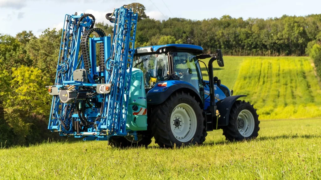 Niebieski ciągnik New Holland z zawieszonym opryskiwaczem marki Berthoud (model Hermes) stoi na zielonej łące. Belka polowa opryskiwacza jest złożona pionowo do transportu, tworząc charakterystyczny kształt litery U za kabiną traktora. W tle widać pagórkowaty teren z lasem i uprawami.