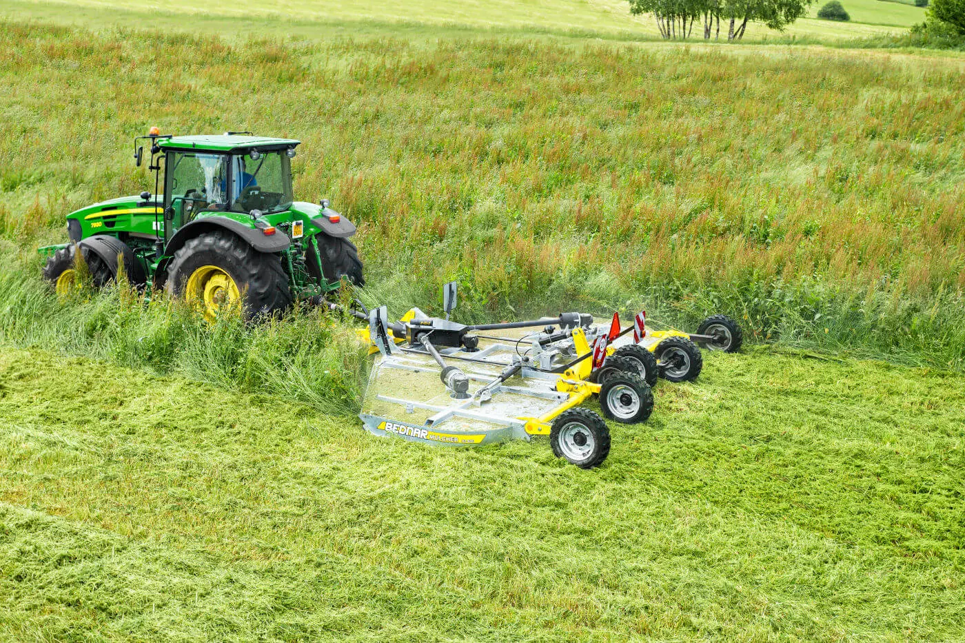 Zielony ciągnik John Deere pracujący z mulczerem Bednar MZ podczas wykaszania wysokiej trawy i nieużytków na zielonym polu.