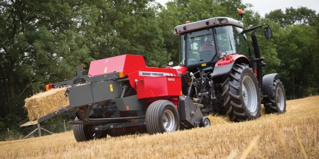 Czerwony ciągnik rolniczy ciągnący małą prasę kostkującą Massey Ferguson 1840 podczas pracy na polu po żniwach. Maszyna wyrzuca ze swojej komory uformowaną, małą, prostokątną belę słomy.