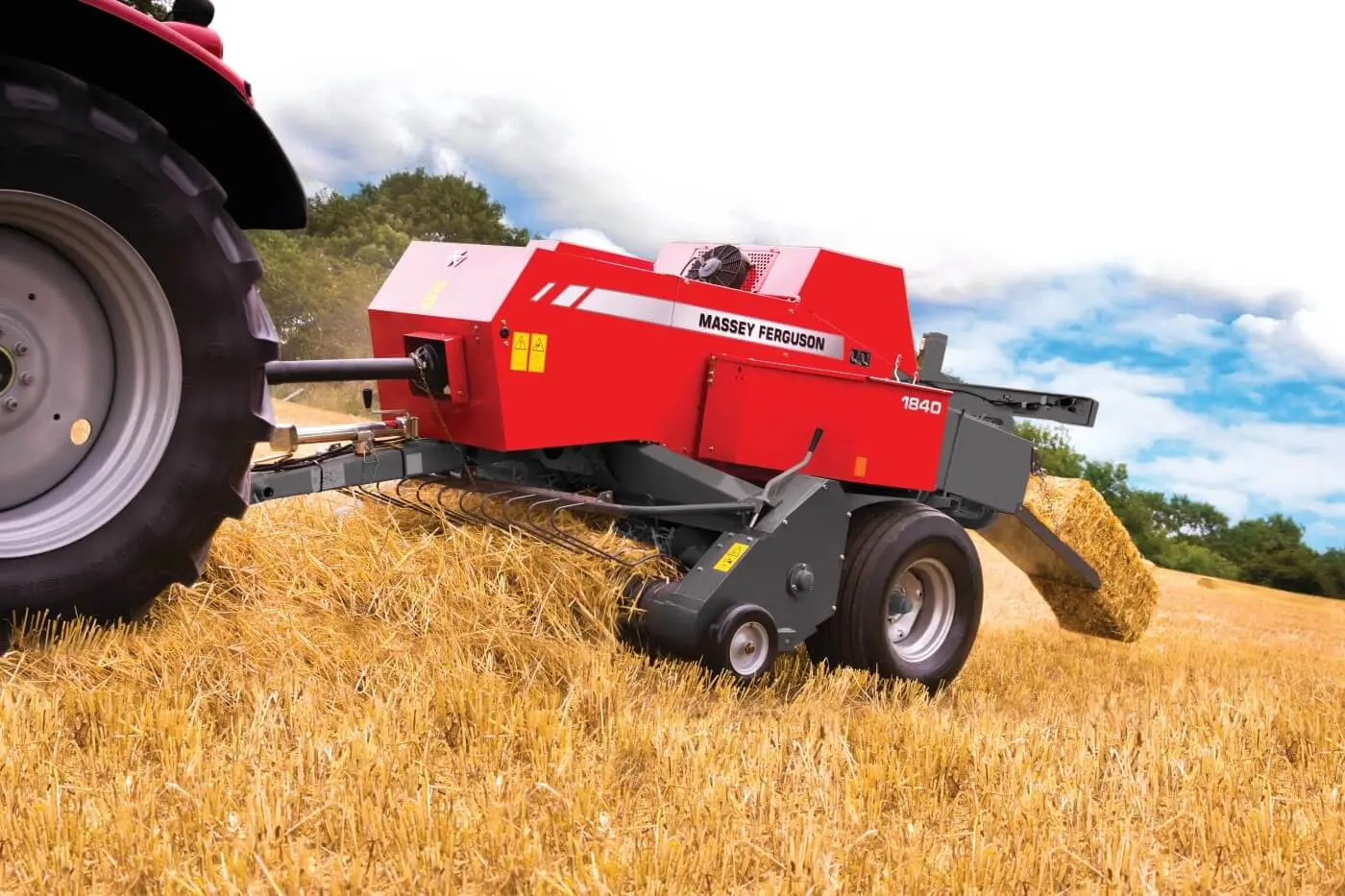 Ujęcie z boku na czerwoną prasę kostkującą Massey Ferguson 1840 podczepioną do ciągnika. Maszyna zbiera słomę z pokosu na ściernisku, a z tyłu urządzenia widać wysuwającą się gotową, małą belę słomy.