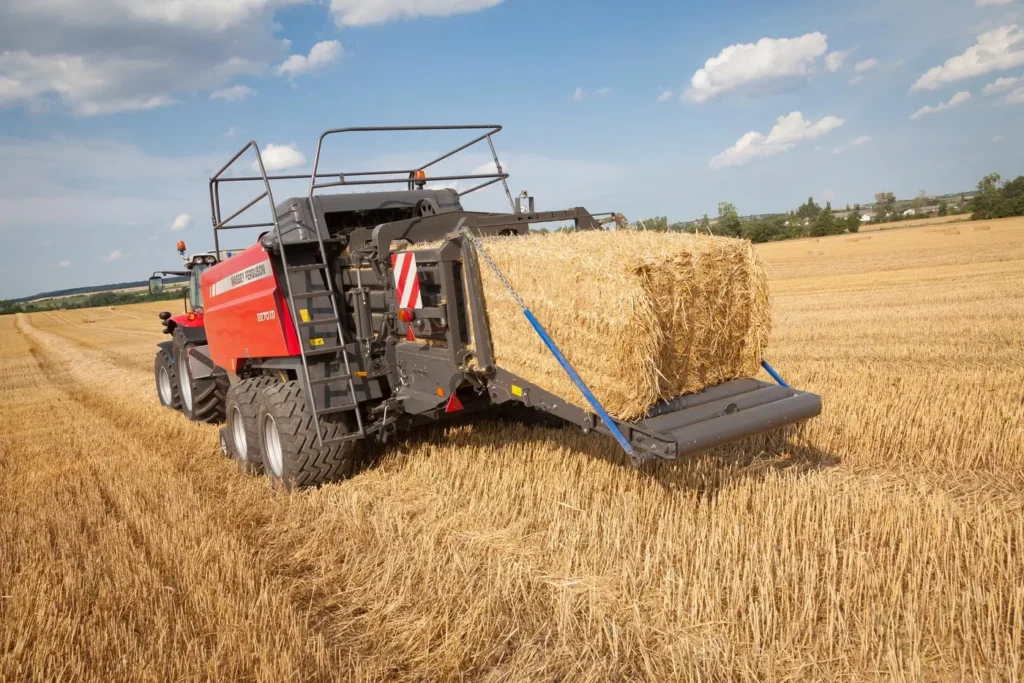 Ciągnik rolniczy z czerwoną, wielkogabarytową prasą kostkującą Massey Ferguson serii 2200 pracujący na ściernisku pod błękitnym niebem z chmurami. Z tyłu maszyny wysuwa się duża, sprasowana, prostokątna bela słomy.