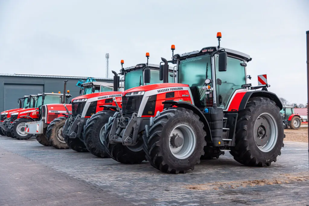 Rząd kilku zaparkowanych obok siebie, czerwonych ciągników rolniczych marki Massey Ferguson, stojących na wybrukowanym placu.