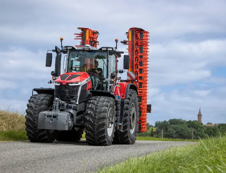 Czerwony ciągnik rolniczy Massey Ferguson ze złożonymi pionowo maszynami rolniczymi, jadący po asfaltowej drodze na tle drzew i zachmurzonego nieba.