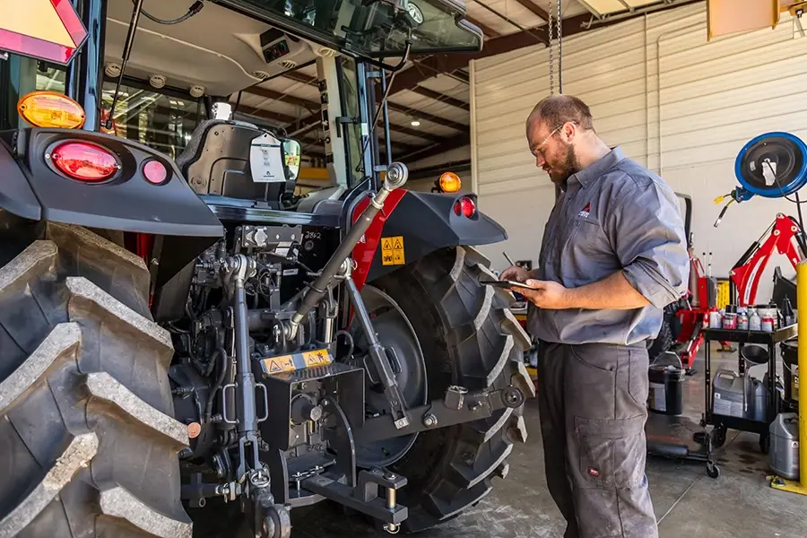 Mechanik w jasno oświetlonym warsztacie, ubrany w koszulę roboczą z logo, sprawdza informacje na tablecie, stojąc przy tylnym układzie zawieszenia i dużym kole czerwonego ciągnika rolniczego.