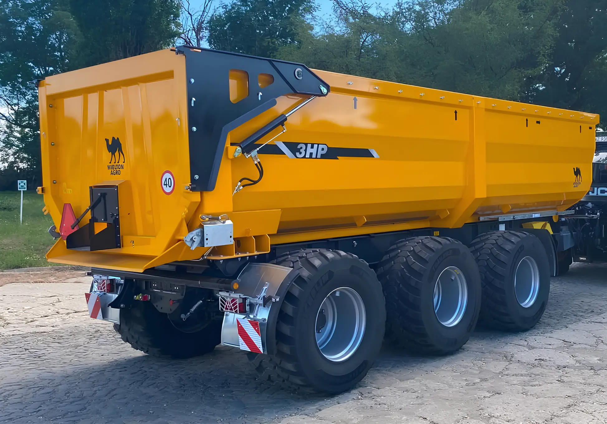 Żółta, trzyosiowa przyczepa rolnicza typu skorupowego (half-pipe) marki Wielton Agro, zaparkowana na brukowanym placu na tle drzew. Na gładkiej burcie bocznej widnieje czarny napis 3HP. Pojazd posiada zawieszenie typu tridem z bardzo dużymi i szerokimi oponami rolniczymi. Z tyłu znajduje się masywna, otwierana hydraulicznie klapa z czarnymi ramionami siłowników, wyposażona w okno zsypowe do zboża, a obok niej naklejka ograniczenia prędkości do 40 km/h oraz logo marki z wielbłądem.