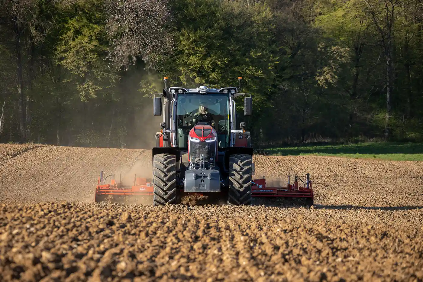 Przednie ujęcie czerwonego ciągnika Massey Ferguson podczas pracy na zaoranym polu, tnącego glebę osprzętem. Wzburzony kurz unosi się za maszyną. W tle gęsty las